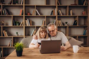 Happy mature 60s husband and wife sit on couch in living room buying things on internet on laptop