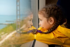 A little girl looks out the window of a train at the sea.